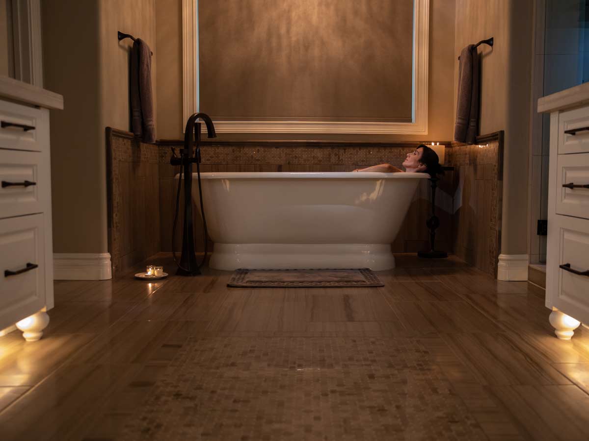 Person relaxing in a bathtub in bathroom with under lit cabinets.